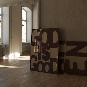 May include: Two rectangular framed wall art prints featuring large, bold, dark brown typography on a light beige background. The primary print displays the text 'GOD IS GOOD' in a stacked layout with the phrase 'all the time' in smaller script.
