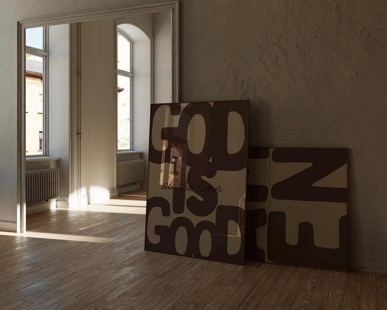 May include: Two rectangular framed wall art prints featuring large, bold, dark brown typography on a light beige background. The primary print displays the text 'GOD IS GOOD' in a stacked layout with the phrase 'all the time' in smaller script.