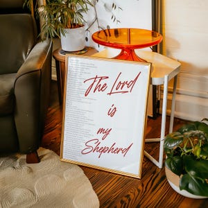 May include: Framed artwork with the text "The Lord is my Shepherd" in red script. The artwork has a gold frame and is displayed on a wooden floor. The image also shows a green armchair, a small side table, and a decorative orange table.