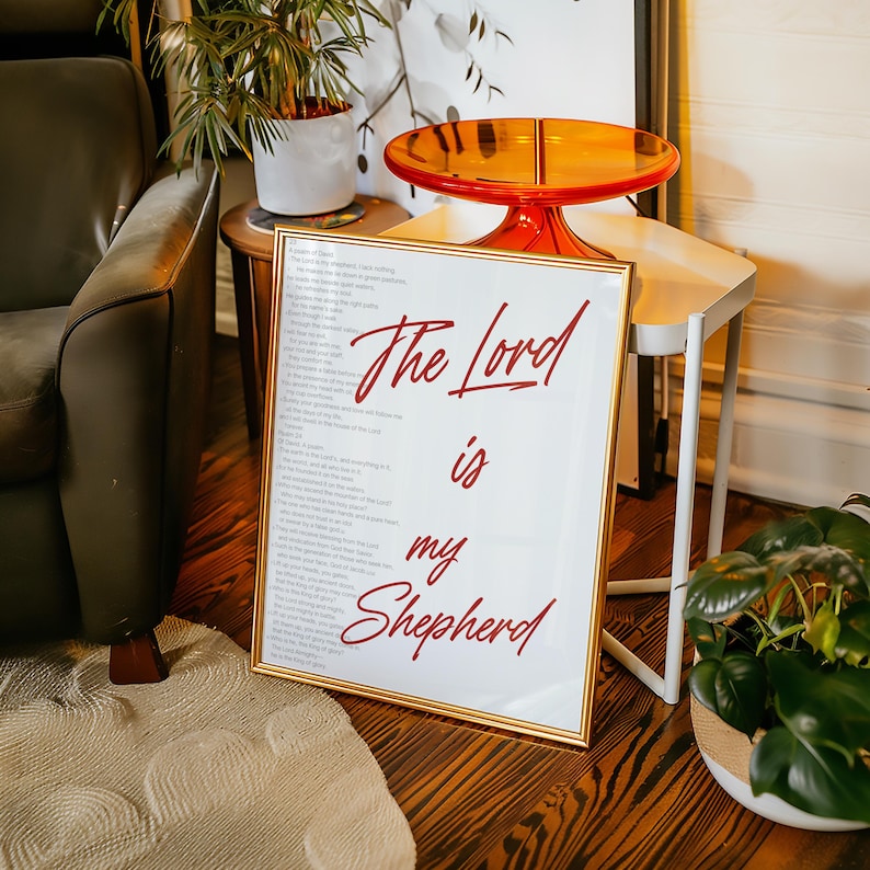 May include: Framed artwork with the text "The Lord is my Shepherd" in red script. The artwork has a gold frame and is displayed on a wooden floor. The image also shows a green armchair, a small side table, and a decorative orange table.