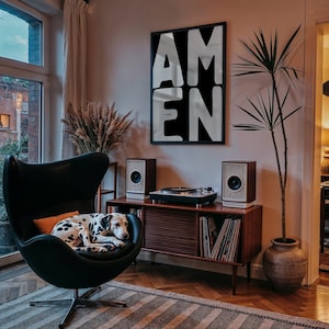 A framed “AMEN” poster hangs above a mid-century console holding speakers and a record player. A cozy lounge chair with a spotted dog curled up rests in the foreground, while plants and warm lighting add character to the room. The dramatic black-and-white design stands out as a modern, faith-inspired décor piece.