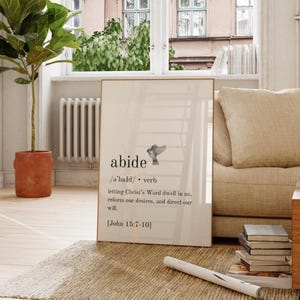 Puede incluir: Impresión enmarcada con la palabra « abide » y su definición, además de una ilustración de un pájaro. El texto negro está sobre un fondo crema. La impresión se muestra en interiores, cerca de una ventana y un sofá.