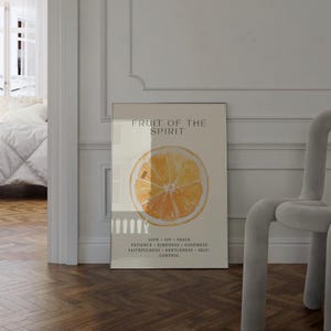 The framed “Fruit of the Spirit” orange watercolor poster stands along a bright hallway wall, with sunlight flowing from an adjacent room. The warm citrus tones and green leaf details contrast beautifully with the soft white walls. The Scripture virtues add an inspiring, faith-filled touch to the minimalist setting.