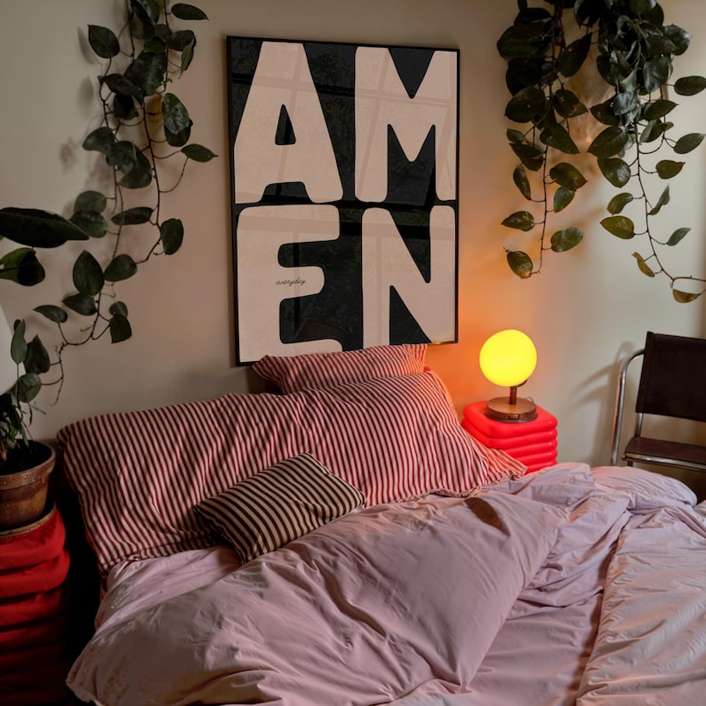 A striking “AMEN” poster hangs above a cozy bed layered with pink bedding and striped pillows. Trailing vine plants frame the wall, while a warm bedside lamp creates a soft ambient glow. The bold black-and-white typography contrasts beautifully with the lush greenery, giving the space a peaceful, faith-centered atmosphere.
