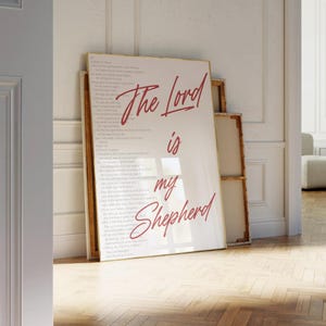 May include: A framed print with the words "The Lord is my Shepherd" in red script. The print is leaning against a wall, with a second, smaller framed print behind it. The background is a light-colored wall and wooden floor.