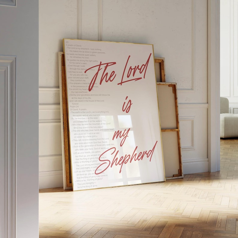 May include: A framed print with the words "The Lord is my Shepherd" in red script. The print is leaning against a wall, with a second, smaller framed print behind it. The background is a light-colored wall and wooden floor.