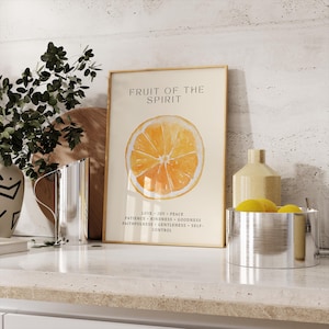 A watercolor orange “Fruit of the Spirit” print leans on a stone countertop next to decorative kitchen jars and a yellow pitcher. Soft neutral tones and gentle light highlight the citrus artwork. The listed Christian virtues complement the peaceful, airy atmosphere of the kitchen corner.