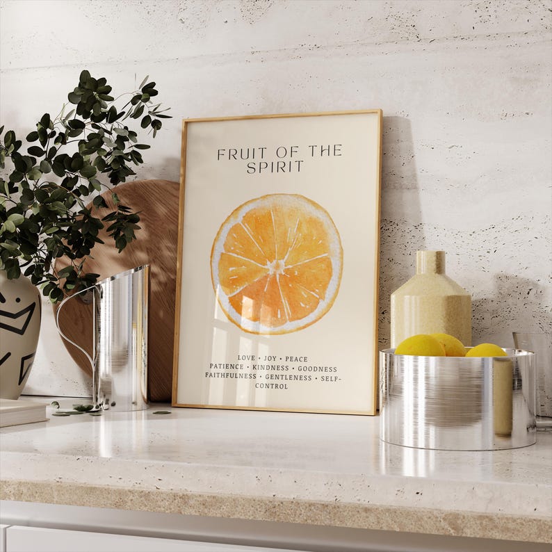 A watercolor orange “Fruit of the Spirit” print leans on a stone countertop next to decorative kitchen jars and a yellow pitcher. Soft neutral tones and gentle light highlight the citrus artwork. The listed Christian virtues complement the peaceful, airy atmosphere of the kitchen corner.