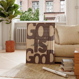 Könnte beinhalten: Ein gerahmter Kunstdruck mit dem Text "GOD IS GOOD all the time" in Braun- und Beigetönen. Das Kunstwerk steht in einem Wohnzimmer, angelehnt an ein beiges Sofa und einen gewebten Teppich, was eine gemütliche Atmosphäre schafft.