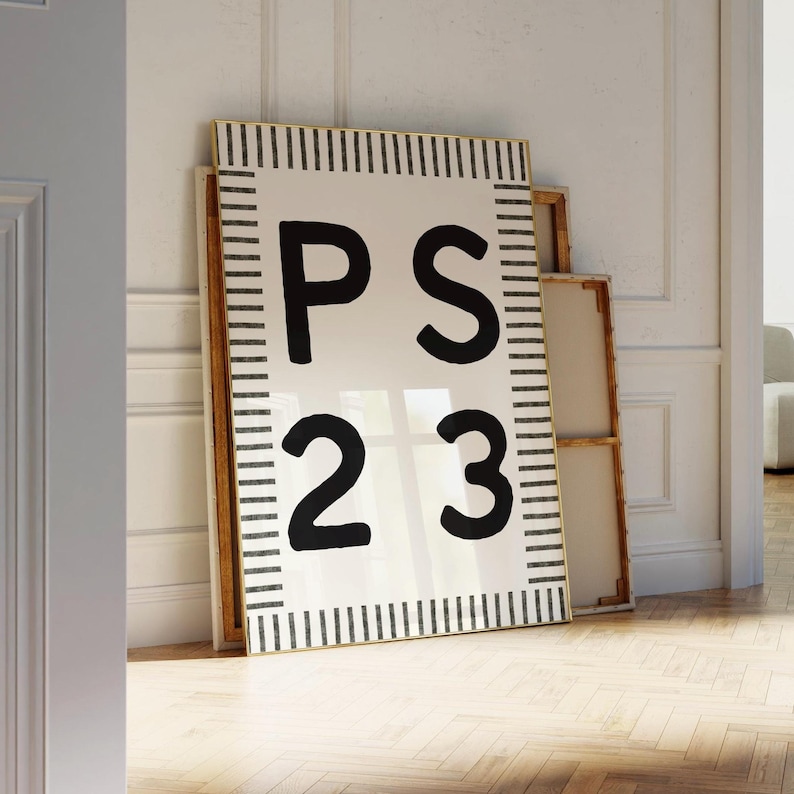 A large Psalm 23 poster with bold black “PS 23” lettering and a modern black-and-white striped border leans against an ornate white wall in a bright, elegant room. Sunlight falls across the herringbone wood floor, highlighting the framed artwork and stacked empty frames around it for a warm, serene aesthetic.