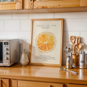 Christian wall art print with watercolor orange design leaning on a kitchen counter. Surrounded by utensils and soft light, the portrait composition blends faith and everyday warmth.