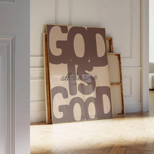 May include: A rectangular wall art print in a thin gold frame featuring the text GOD IS GOOD in large, bold, dark brown block letters against a light beige background, with the phrase all the time written in a smaller, dark brown cursive script.