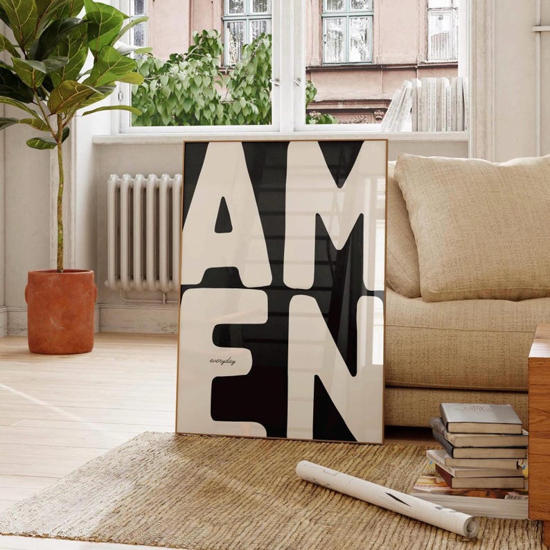 A bold black-and-white “AMEN” poster leans against a beige couch in a bright living room. Sunlight fills the space through large windows, highlighting a woven rug, stacked books, and a tall plant. The clean, modern interior enhances the strong typography, making the faith-inspired printable artwork stand out as a powerful décor piece.