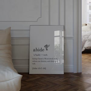 Op de afbeelding: Een ingelijste print met het woord "abide" en de definitie, plus een bijbelvers. Een kleine illustratie van een vogel staat boven de definitie. De print staat tegen een witte muur.