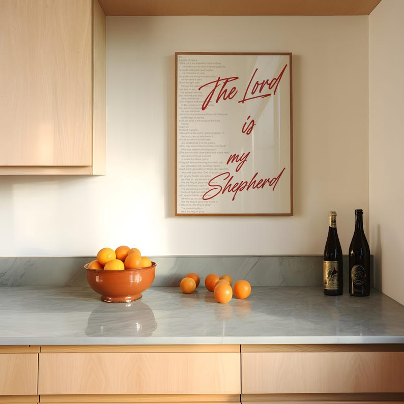 May include: A kitchen scene featuring a framed print with the text "The Lord is my Shepherd" in red script. A terracotta bowl filled with oranges sits on a gray countertop, with additional oranges scattered around. Two wine bottles are also visible.