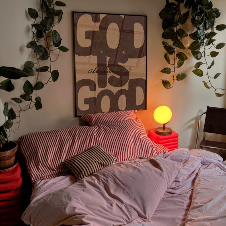 May include: A rectangular wall art print in a black frame featuring the text "GOD IS GOOD" in large, bold, dark brown letters against a light beige background. The print is displayed above a bed with striped bedding and a solid light pink duvet.