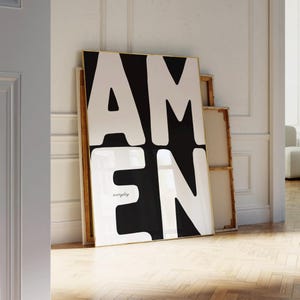 A large black-and-white “AMEN” poster leans against a textured white wall beside stacked wooden frames. Warm sunlight spreads across the herringbone floor, casting natural shadows that highlight the clean architectural details. The minimalist, elegant setting complements the strong typographic design of this faith-inspired printable artwork.