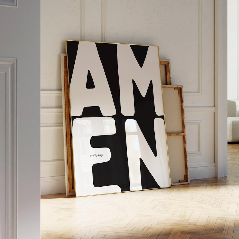 A large black-and-white “AMEN” poster leans against a textured white wall beside stacked wooden frames. Warm sunlight spreads across the herringbone floor, casting natural shadows that highlight the clean architectural details. The minimalist, elegant setting complements the strong typographic design of this faith-inspired printable artwork.