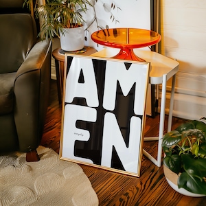 A bold “AMEN” poster leans on a small wooden table decorated with a bright orange tray and surrounding greenery. Soft daylight warms the wall and herringbone floor, creating a cozy and stylish vignette. The strong black-and-white lettering becomes the focal point, adding a modern faith-centered touch to the interior.