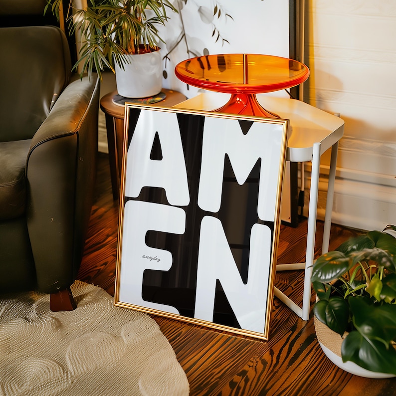 A bold “AMEN” poster leans on a small wooden table decorated with a bright orange tray and surrounding greenery. Soft daylight warms the wall and herringbone floor, creating a cozy and stylish vignette. The strong black-and-white lettering becomes the focal point, adding a modern faith-centered touch to the interior.