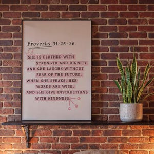 Op de afbeelding: Een rechthoekige wandkunstprint in een zwarte lijst met de tekst Proverbs 31:25-26, She is clothed with strength and dignity and she laughs without fear of the future. When she speaks, her words are wise, and she give instructions with kindness.