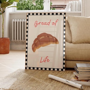 May include: Framed art print featuring a watercolor illustration of a croissant. The words "Bread of Life" are written in a cursive font. The frame has a black and white checkered border. The print is displayed on a rug.