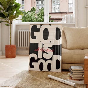 May include: A framed art print featuring the words "GOD IS GOOD all the time" in bold black and white. The phrase "all the time" is in red cursive. The print is displayed on a rug in a living room, with a neutral colour scheme.