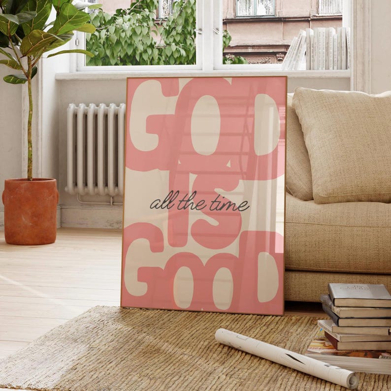 May include: A framed art print with the words "GOD IS GOOD all the time" in pink over a cream background. The print is leaning against a beige sofa and a stack of books. A potted plant and a window are in the background.