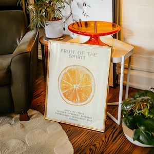 The “Fruit of the Spirit” orange poster leans against a small wooden side table topped with a translucent orange tray. Plants and warm lighting surround the scene, echoing the citrus tones of the watercolor artwork. The soft cream background and Scripture virtues create a comforting, uplifting aesthetic.
