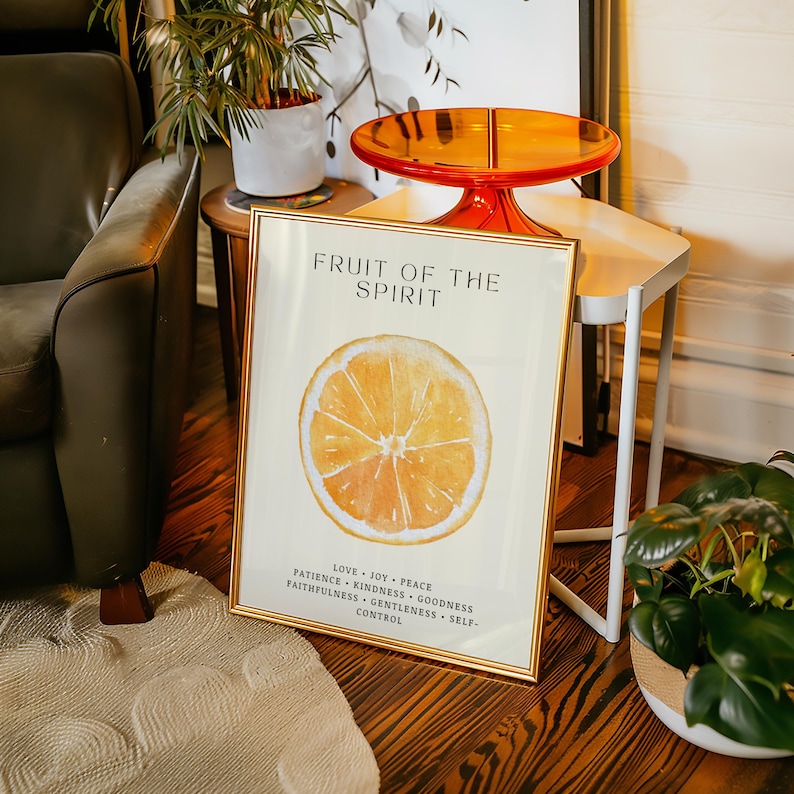 The “Fruit of the Spirit” orange poster leans against a small wooden side table topped with a translucent orange tray. Plants and warm lighting surround the scene, echoing the citrus tones of the watercolor artwork. The soft cream background and Scripture virtues create a comforting, uplifting aesthetic.