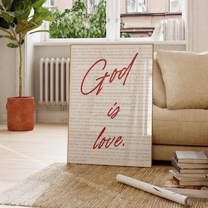 May include: A framed canvas print featuring the words "God is love" in red script, set against a backdrop of printed text. The print is displayed in a bright room with a plant and a sofa.