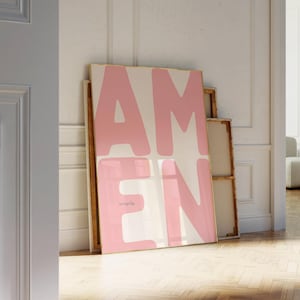 Puede incluir: Una impresión artística enmarcada con la palabra "AMEN" en letras grandes y rosas sobre un fondo blanquecino. La palabra "everyday" está impresa en una fuente más pequeña debajo de la letra "E". La obra de arte está apoyada contra una pared blanca.