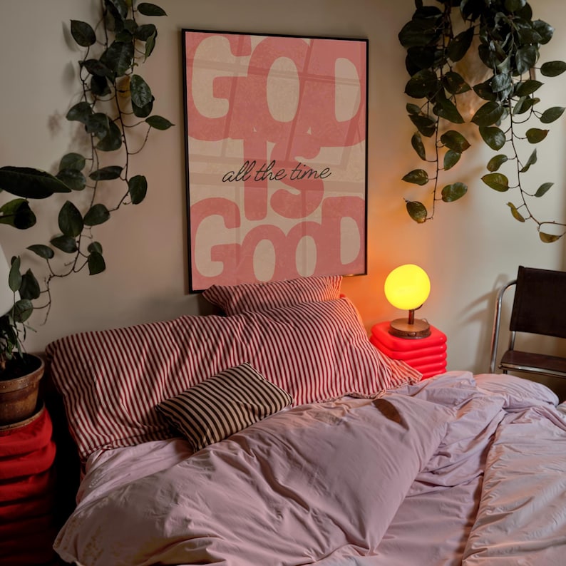 May include: A bedroom scene featuring a bed with pink bedding and striped pillows. A framed poster with the words "GOD IS GOOD all the time" hangs above the bed. A small lamp with a yellow globe sits on a red cube, and greenery hangs from the ceiling.