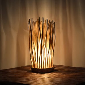 May include: A decorative table lamp featuring a unique design of vertical, light-colored wooden branches. The branches are arranged around a light source, creating a warm, ambient glow. The lamp sits on a wooden base, adding to its natural aesthetic.
