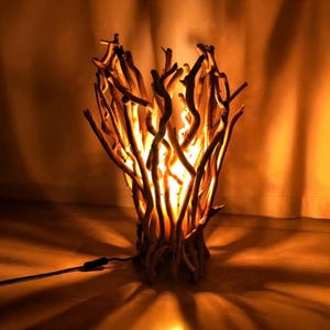 Puede incluir: Lámpara de mesa artesanal hecha de madera flotante. La lámpara presenta un grupo de ramas de madera entrelazadas, creando un brillo cálido. La base de la lámpara también es de madera y tiene un cable de alimentación negro. La lámpara mide unos 30 cm de alto.