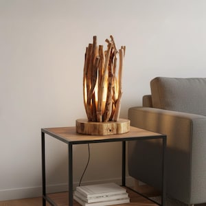 Puede incluir: Una lámpara de mesa rústica de madera. La lámpara tiene una base de disco de madera, con numerosos palos de madera dispuestos alrededor de una fuente de luz central. La lámpara está colocada sobre una mesa auxiliar de madera con marco de metal negro.