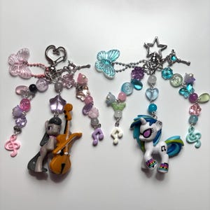 May include: Two decorative keychains featuring pony figurines. The left keychain has a gray pony with a cello, pink butterfly, and music note charms. The right keychain has a white pony with rainbow accents, a blue butterfly, and music note charms.