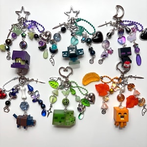 May include: A collection of six decorative keychains featuring various Minecraft-inspired character figures, including a purple block, a green slime, and an orange ghast, adorned with colorful beads, charms, stars, hearts, and metal sword accents.