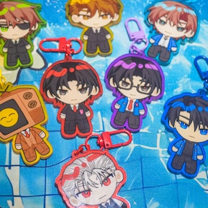 May include: A collection of acrylic keychains featuring various anime-style characters with diverse hair colours, outfits, and accessories, including one character with a television head displaying a yellow smiley face, all attached to colourful plastic clasps.
