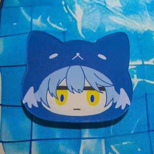 May include: A flat acrylic charm featuring a stylized character head with light blue hair and bright yellow eyes, wearing a dark blue cat-shaped hood with small white ears and a simple white cat nose and mouth graphic printed on the top edge.