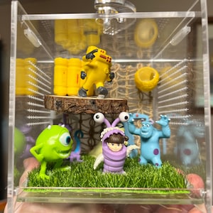 Puede incluir: Una vitrina de acrílico transparente con figuras en miniatura de la película Monstruos, S.A. La escena incluye a Mike Wazowski, Sulley, Boo y otros personajes, sobre césped artificial. También hay una figura amarilla de traje de protección.