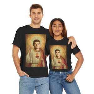 May include: Black t-shirts featuring a portrait of a person with a halo, a red heart, and a gold background. The person is wearing a white robe and a red shawl. The image is a parody of religious art.