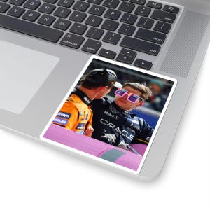 May include: A sticker featuring two people in racing attire. One person wears a black uniform with "ORACLE" and "Mobil 1" logos, and pink-framed sunglasses. The other person wears an orange uniform. The sticker is on a laptop.