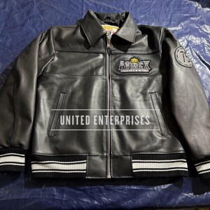 May include: Black leather jacket with a zip-up front, collared neckline, and ribbed cuffs and hem with white stripes. Features the "AVIREX" logo patch on the chest and a "75" patch on the sleeve. The text "UNITED ENTERPRISES" is printed across the front.