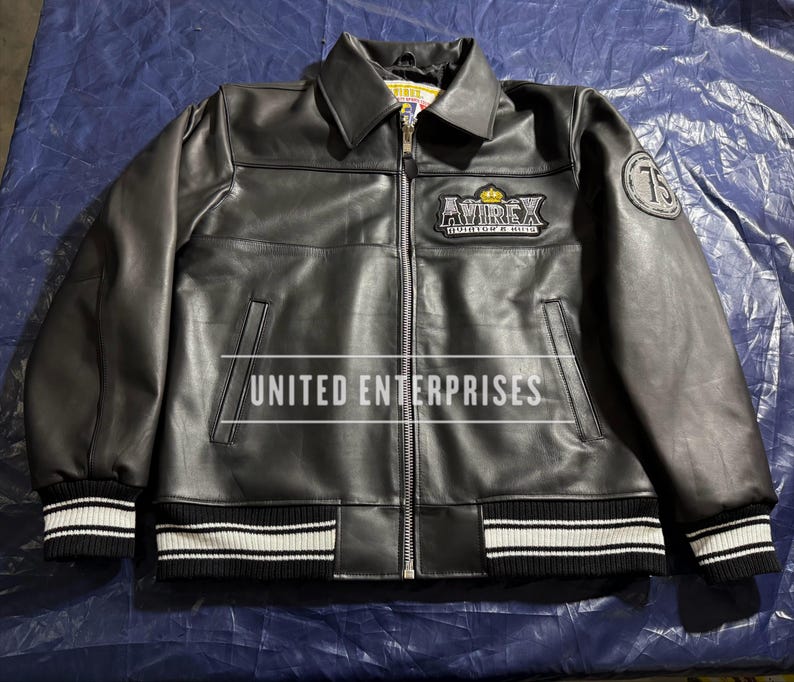 May include: Black leather jacket with a zip-up front, collared neckline, and ribbed cuffs and hem with white stripes. Features the "AVIREX" logo patch on the chest and a "75" patch on the sleeve. The text "UNITED ENTERPRISES" is printed across the front.