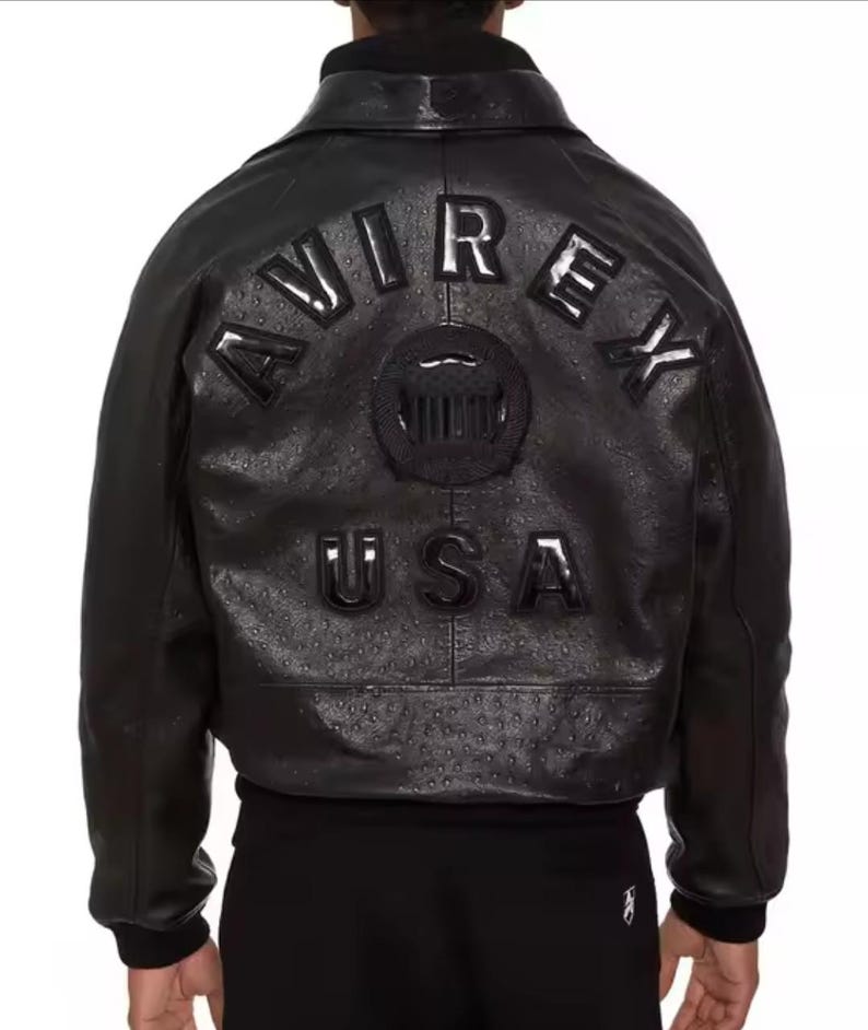 Op de afbeelding: Zwarte leren jas met het woord "AVIREX" in verhoogde letters op de rug, samen met een cirkelvormig embleem en het woord "USA". De jas heeft een getextureerde afwerking en een bomberjack-ontwerp.