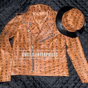 Puede incluir: Chaqueta de cuero marrón y sombrero de cubo a juego. La chaqueta presenta un estilo motero clásico con cremallera diagonal y múltiples bolsillos. Ambos artículos están cubiertos con un patrón repetido de logotipos negros. El texto "UNITED ENTERPRISES" es visible.