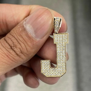 May include: A gold-colored pendant in the shape of the letter "J", encrusted with numerous small, sparkling gemstones. The pendant is held by a hand, showcasing its detailed design and craftsmanship. The jewelry piece is a personalized accessory.