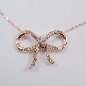 Puede incluir: Collar con colgante de lazo en oro rosa con una cadena delicada. El lazo está incrustado con pequeñas piedras claras y brillantes. El collar se muestra sobre un fondo blanco.