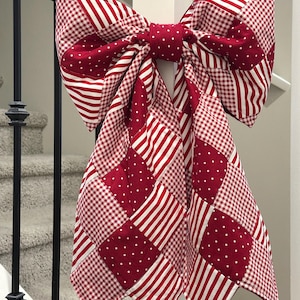 May include: A large, decorative bow in red and white, attached to a white post. The bow is made of fabric with various patterns, including stripes, polka dots, and gingham. The bow is a festive decoration, possibly for a holiday or special occasion.